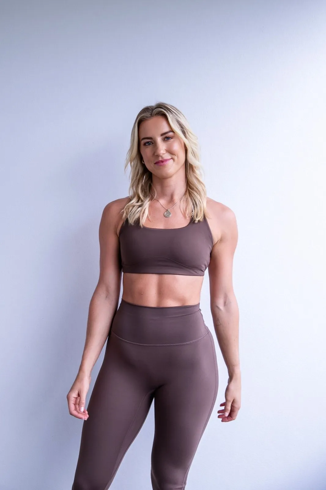 Charlotte Hazelwood, strength and mobility coach, standing against a white wall in brown training wear