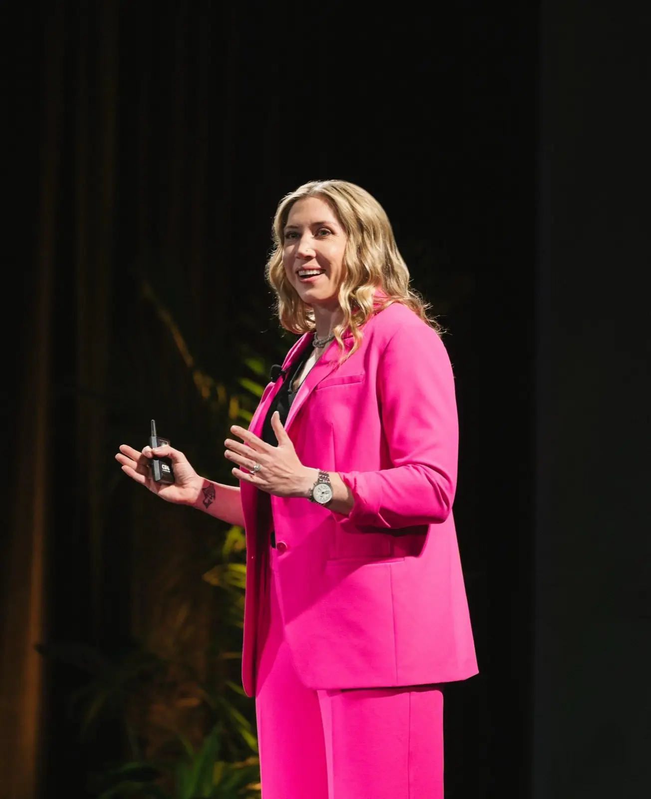Michelle “Mace” Curran speaking on stage in a pink suit, holding a slide clicker